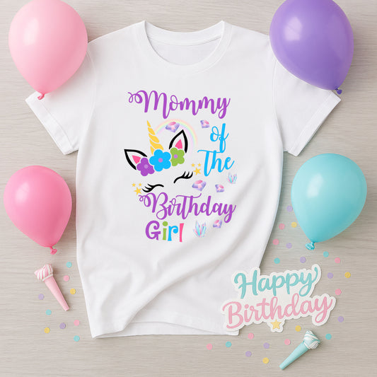 White t-shirt with 'Mommy of the Birthday Girl' text and unicorn design, surrounded by balloons and confetti on a wooden surface.